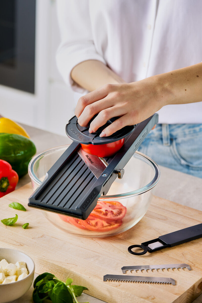 Home cooking using mandolin slicer for cutting tomato with finger safety guard for safe and even slicing of tomato into thin, uniform pieces.