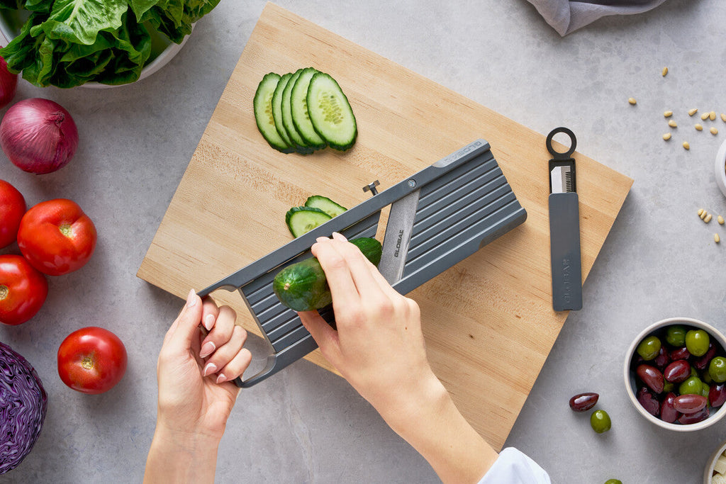 Cooking enthusiast using easy to use Global Japanese mandoline for cutting cucumber into thin, even slices on wooden cutting board for healthy meal prep.