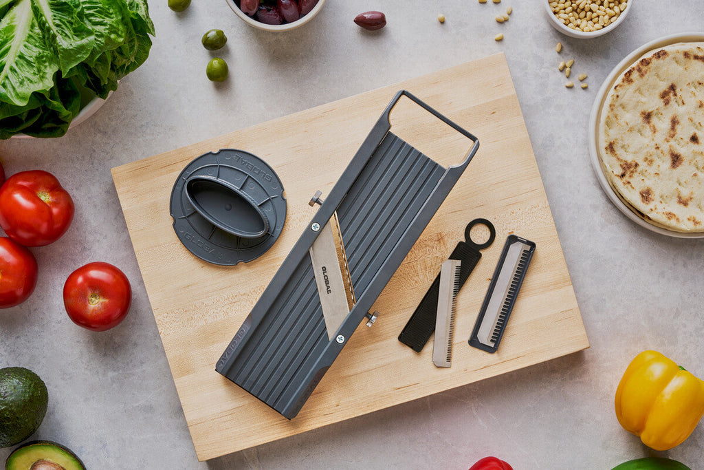 Globals 6.4cm mandolin veggie cutter on a wooden cutting board with vegetables around, 3 mandolin slicer blade attachments and a hand guard for a safe mandoline slicer.