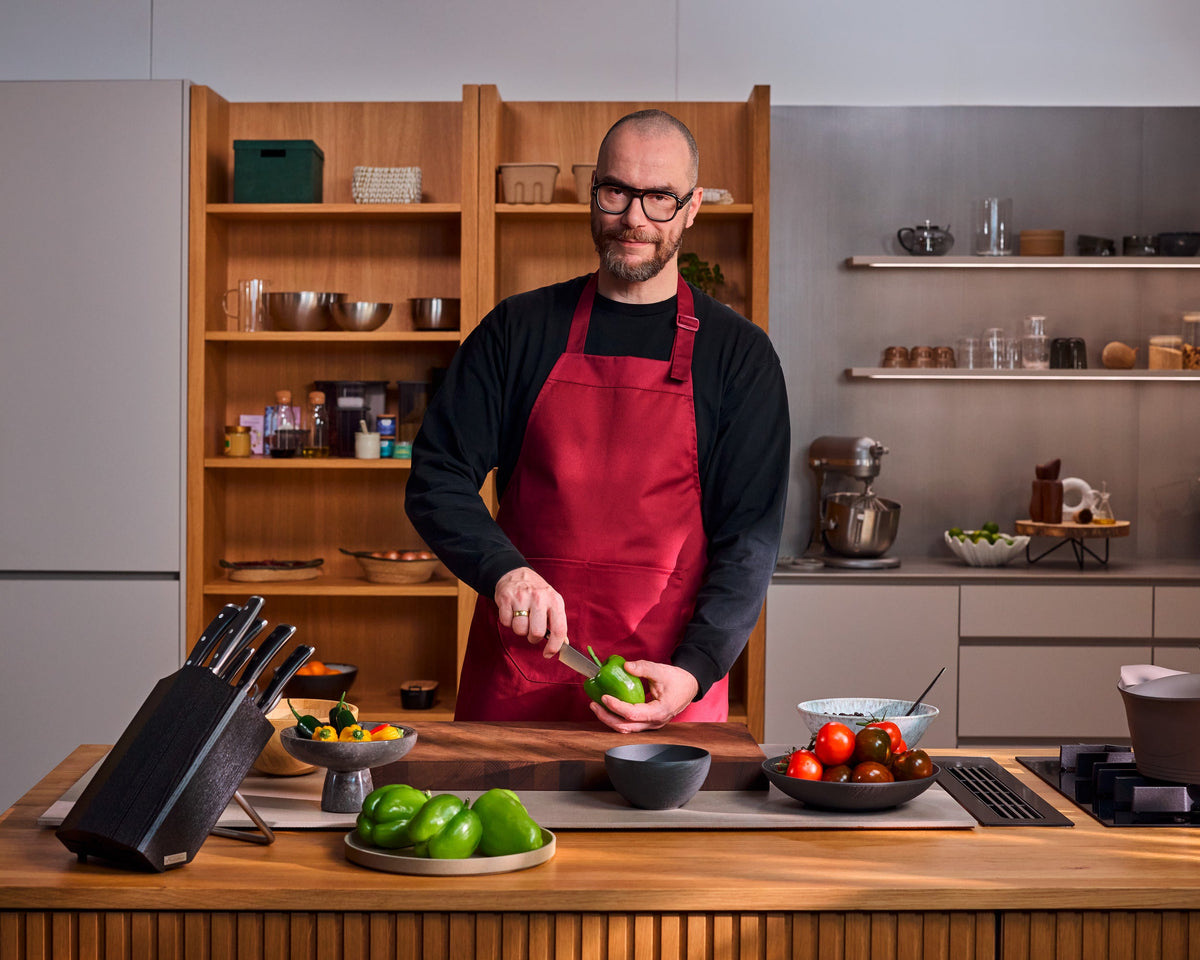 Man in a red apron slicing a green bell pepper with the Classic Ikon Asian Utility Knife 12 cm | 4 1/2 inch in a modern kitchen setting.