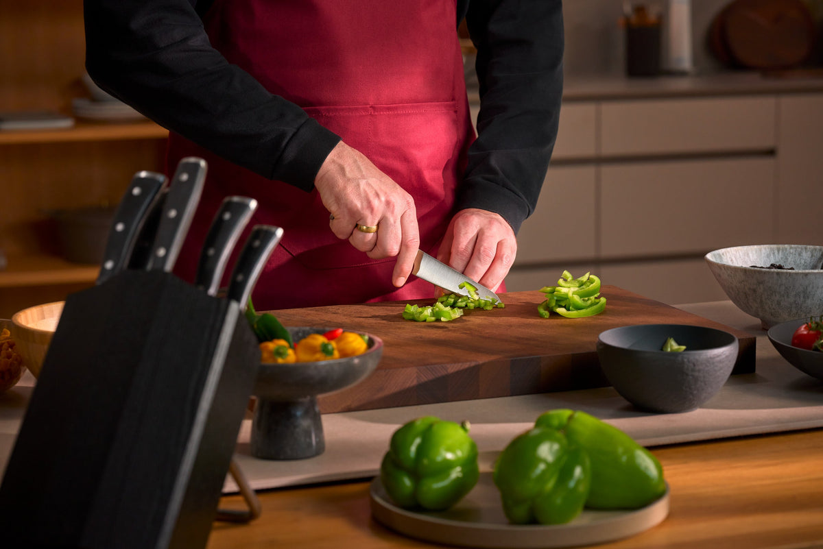 Classic Ikon Asian Utility Knife 12 cm | 4 1/2 inch slicing green bell pepper on a wooden cutting board with a set of knives in the background