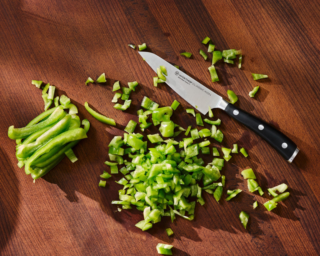 Classic Ikon Asian Utility Knife 12 cm | 4 1/2 inch with a forged stainless steel blade and full tang black handle chopping fresh green bell pepper on a wood surface