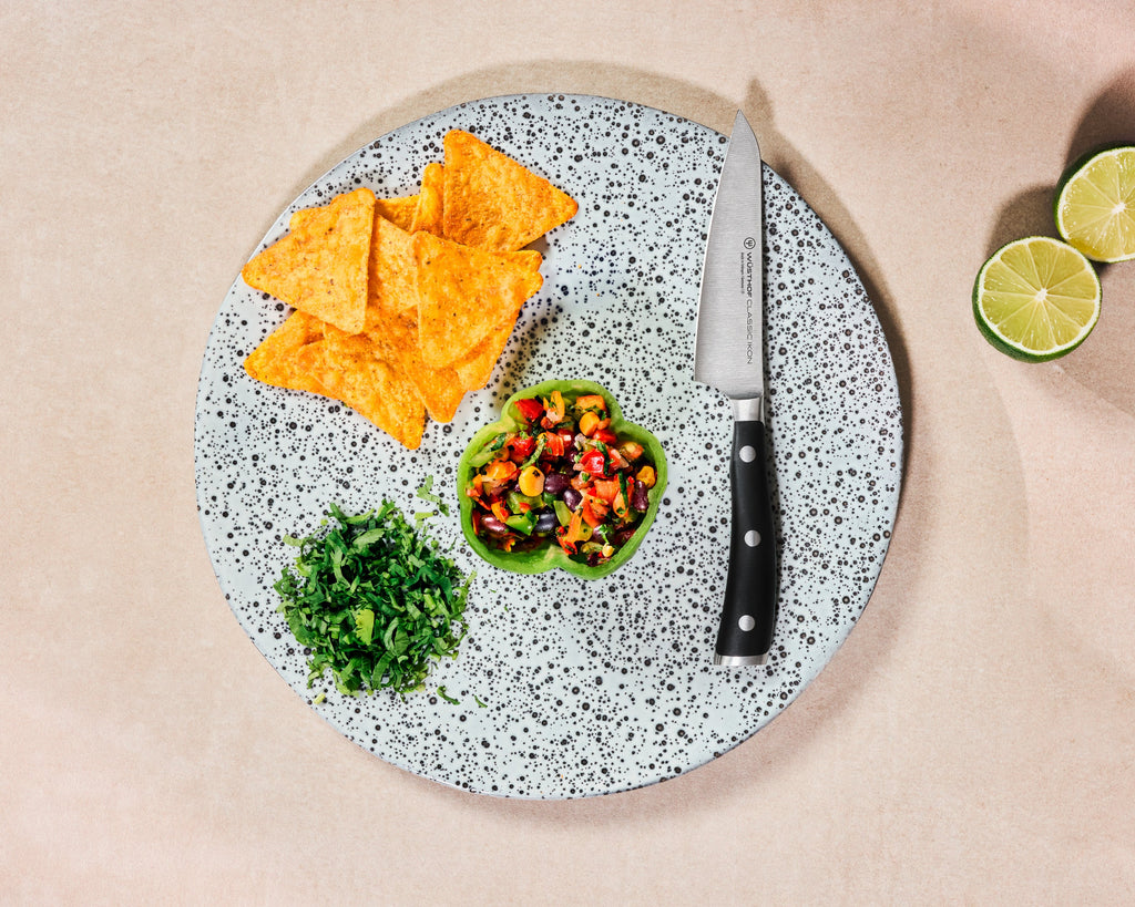 Classic Ikon Asian Utility Knife 12 cm | 4 1/2 inch with a sharp stainless steel blade and a sturdy black handle, displayed on a speckled plate with chopped herbs and salsa.