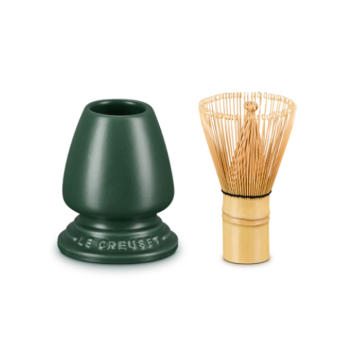 A Le Creuset green bamboo whisk holder next to a traditional Japanese bamboo chashaku whisk with gold-toned tines, set against a solid white background.