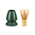 A Le Creuset green bamboo whisk holder next to a traditional Japanese bamboo chashaku whisk with gold-toned tines, set against a solid white background.